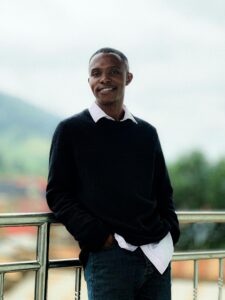A man in casual attire smiles warmly while leaning on a railing outdoors.