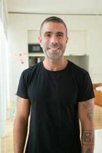 Casually dressed man smiling, standing in a bright modern living room with tattoos visible on his arm.